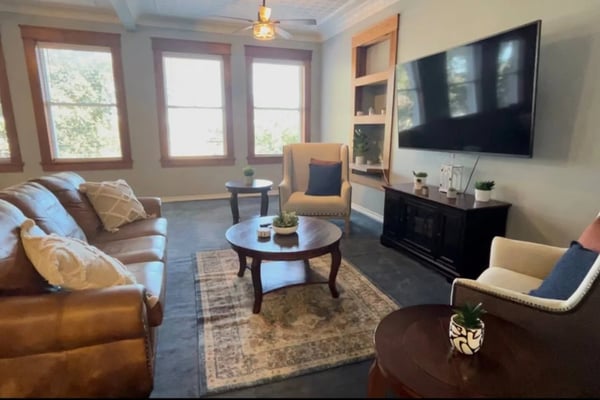 Bright living room with leather sofa, armchairs, round coffee table, wall-mounted TV, and three large windows with wood frames overlooking greenery.