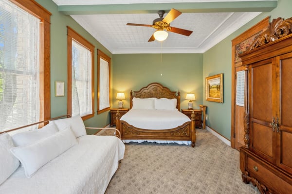Bedroom with sage green walls, carved wooden bed, white sofa, ceiling fan, and wood-framed windows